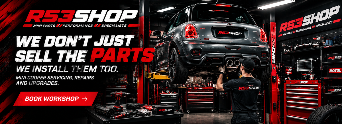 A dark gray Mini Cooper is raised on a hydraulic lift inside R53Shop workshop, with a mechanic working underneath. The image features R53Shop and Mini cooper Parts.